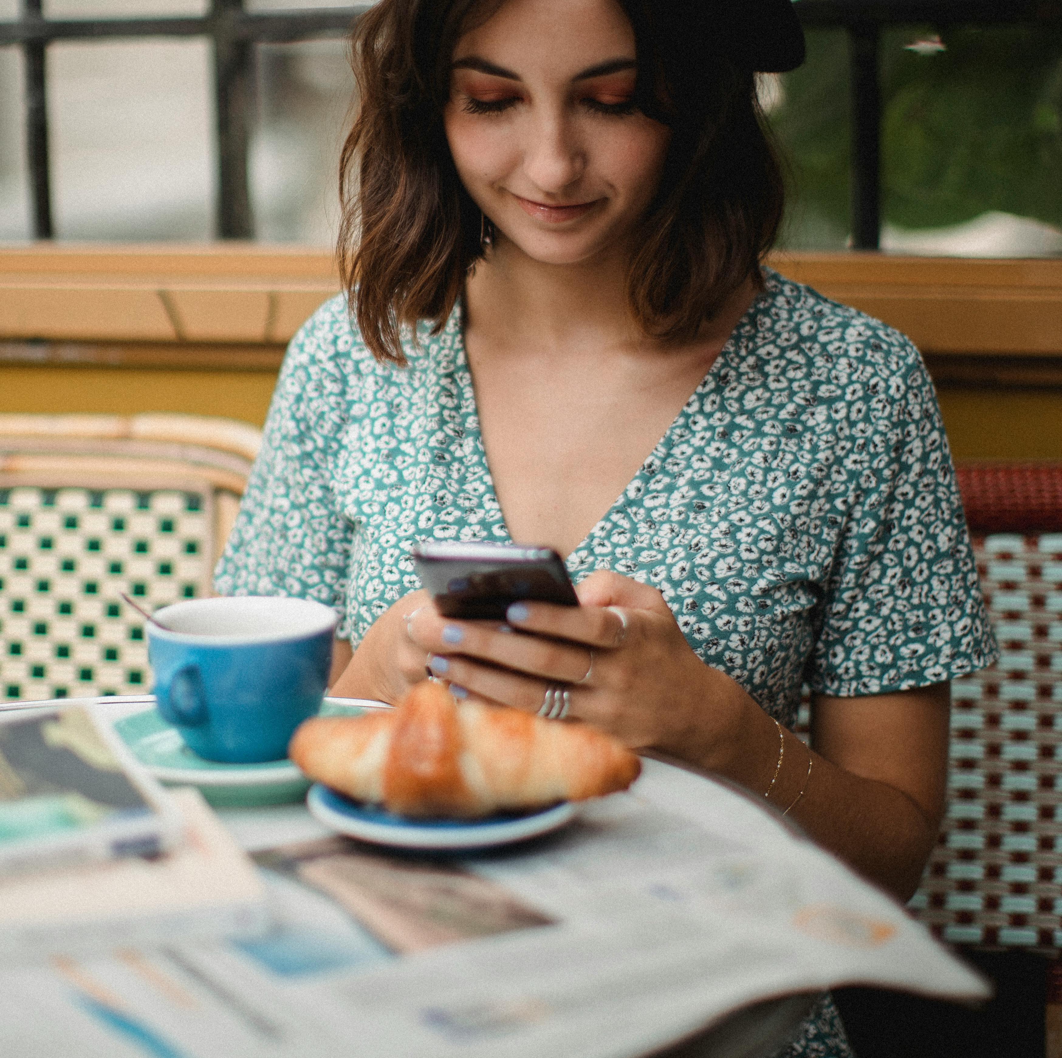 Woman smiling at her phone at a café table with a croissant and coffee